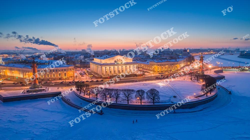На фотографии запечатлена Стрелка Васильевского острова в зимнее утро. Видны Ростральные колонны и здание Биржи, подсвеченные огнями. Залив покрыт льдом, а снег подчёркивает холодную красоту Санкт-Петербурга. Рассветные краски украшают небо, добавляя тепла в зимний пейзаж.